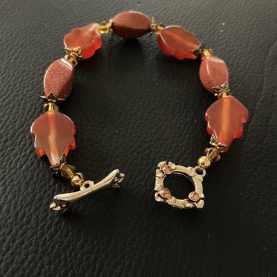 Sale Photo Thumbnail #131: Beaded bracelet featuring carved leaf-shaped translucent orange beads alternating with sparkling goldstone oval beads and small amber-tone spacer accents. Finished with a detailed toggle clasp embellished with small rhinestones. Shows light pre-owned wear