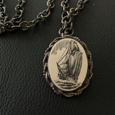 Sale Photo Thumbnail #134: A vintage oval pendant featuring a scrimshaw-style illustration of a sailing ship at sea, set within a twisted metal frame. The piece features a pin back, allowing it to be worn as a brooch, as well as a loop attached to a chunky metal link chain. The pen