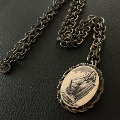 Sale Photo Thumbnail #135: A vintage oval pendant featuring a scrimshaw-style illustration of a sailing ship at sea, set within a twisted metal frame. The piece features a pin back, allowing it to be worn as a brooch, as well as a loop attached to a chunky metal link chain. The pen
