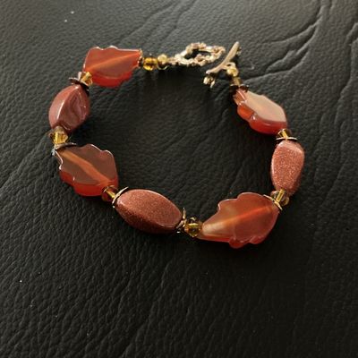 Sale Photo Thumbnail #132: Beaded bracelet featuring carved leaf-shaped translucent orange beads alternating with sparkling goldstone oval beads and small amber-tone spacer accents. Finished with a detailed toggle clasp embellished with small rhinestones. Shows light pre-owned wear