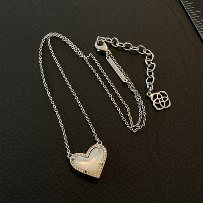 Sale Photo Thumbnail #172: A Kendra Scott silver-tone necklace featuring a heart-shaped mother of pearl (iridescent stone) pendant with a textured or pave-accented border. Includes an adjustable extender chain with the signature Kendra Scott logo charm and branded metal tag near th