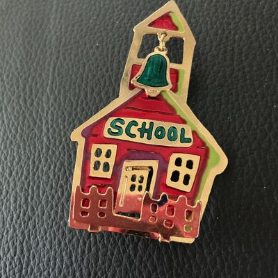 Sale Photo Thumbnail #150: A gold-tone and multicolored enamel brooch shaped like a red schoolhouse with a fence and a dangling green bell charm inside the belfry. Features a pin-back closure with standard rollover clasp. Marked on the back with a maker's mark showing two hands. Me