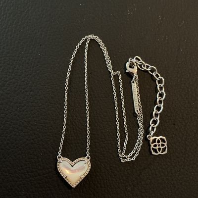 Sale Photo Thumbnail #171: A Kendra Scott silver-tone necklace featuring a heart-shaped mother of pearl (iridescent stone) pendant with a textured or pave-accented border. Includes an adjustable extender chain with the signature Kendra Scott logo charm and branded metal tag near th