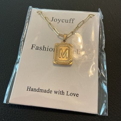 Sale Photo Thumbnail #190: Joycuff gold-tone fashion necklace featuring a rectangular pendant with the initial 'M' on a paperclip style chain. New in original plastic packaging with card insert.