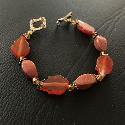 Sale Photo Thumbnail #130: Beaded bracelet featuring carved leaf-shaped translucent orange beads alternating with sparkling goldstone oval beads and small amber-tone spacer accents. Finished with a detailed toggle clasp embellished with small rhinestones. Shows light pre-owned wear