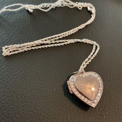 Sale Photo Thumbnail #224: A silver-tone rope chain necklace featuring a heart-shaped locket pendant. The front of the locket has a glittery enamel-style inlay bordered by clear rhinestones, and opens to reveal two picture slots. Includes an adjustable extender chain with a lobster