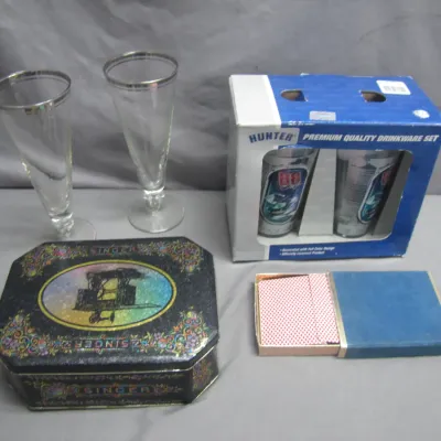 Sale Photo Thumbnail #95: Assortment of collectibles including two silver-rimmed pilsner glasses, a boxed Hunter NASCAR drinkware set, a vintage decorative Singer sewing machine tin, and a boxed deck of playing cards.