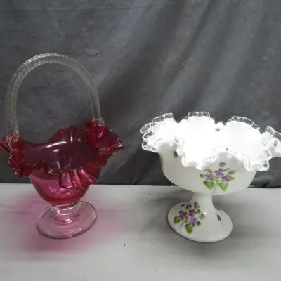 Sale Photo Thumbnail #11: Fenton art glass footed basket in cranberry glass with a clear applied handle alongside a hand-painted white milk glass compote bowl featuring floral details and a ruffled rim.