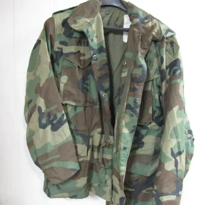 Sale Photo Thumbnail #68: US Military M-65 woodland camouflage field jacket hanging on a black hanger, featuring front utility pockets and shoulder epaulets.