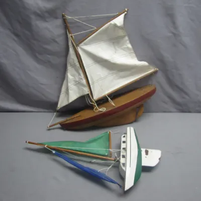 Sale Photo Thumbnail #86: Two vintage wooden toy sailboats, including a gaff-rigged model with white cloth sails on a wooden stand and a painted green and white model lying flat.