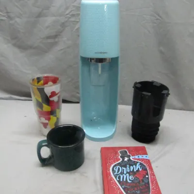 Sale Photo Thumbnail #301: Light blue SodaStream sparkling water maker shown alongside a Maryland flag tumbler, ceramic mug, car cup holder adapter, and a 'Drink Me' cocktail book.