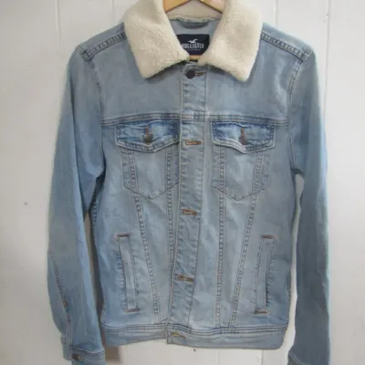 Sale Photo Thumbnail #223: Hollister light-wash denim trucker jacket with a fleece-lined collar, displayed on a wooden hanger.