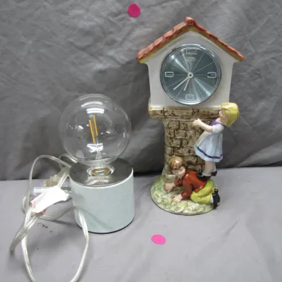 Sale Photo Thumbnail #118: Vintage ceramic clock featuring children playing by a stone tower alongside a small modern accent lamp with a filament bulb.