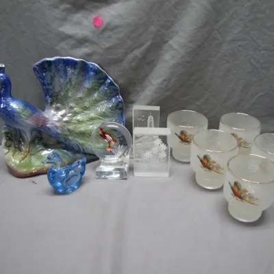 Sale Photo Thumbnail #184: Collection of decorative glassware and figurines, featuring a iridescent ceramic peacock, glass duck paperweights, etched glass blocks, and a set of frosted lowball glasses with bird illustrations.