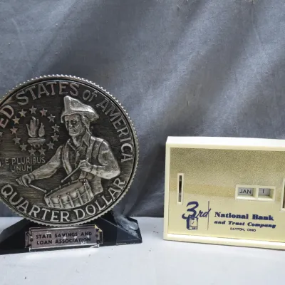 Sale Photo Thumbnail #13: Pair of vintage bank promotional desktop collectibles, including a Bicentennial quarter-style coin bank from State Savings and Loan and a 3rd National Bank flip calendar.