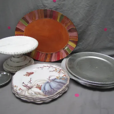 Sale Photo Thumbnail #189: Collection of serving items including a distressed white pedestal cake stand, autumn-themed ceramic plates, metal chargers, and a large striped oval platter.