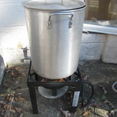 Sale Photo Thumbnail #161: Large aluminum stock pot with lid and spigot valve, mounted on a black metal propane outdoor burner stand.
