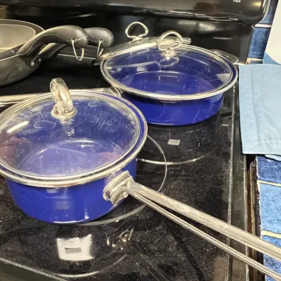 Sale Photo Thumbnail #25: Cobalt blue enamel cookware including a saucepan and a skillet, both featuring clear glass lids and stainless steel loop handles.