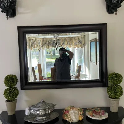 Sale Photo Thumbnail #39: Large dark-framed wall mirror hanging above a half-moon console table with decorative tableware, flanked by black ornate sconces and artificial topiary plants.