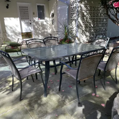 Sale Photo Thumbnail #23: Outdoor patio dining set featuring a rectangular glass-top table with a dark metal frame and six matching woven wicker-backed armchairs.