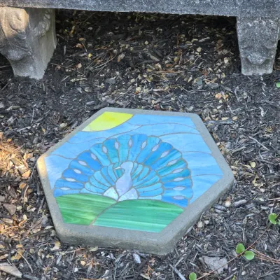 Sale Photo Thumbnail #51: Hexagonal concrete garden stepping stone featuring a colorful mosaic peacock design, displayed outdoors near a stone bench.