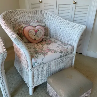 Sale Photo Thumbnail #56: Vintage white painted wicker barrel chair with floral upholstered seat cushion, accompanied by decorative heart pillow and a fringed footstool.