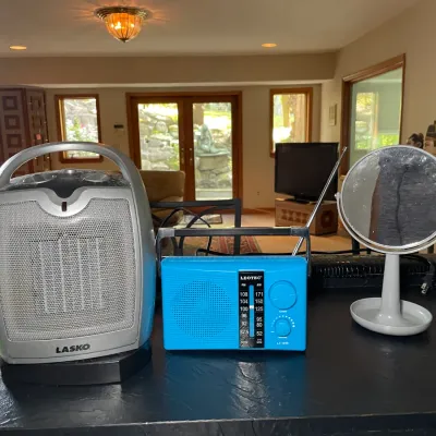 Sale Photo Thumbnail #8: Collection of small household electronics, including a silver Lasko ceramic space heater, a blue Leotec portable FM/AM radio with telescopic antenna, and a small round vanity mirror.