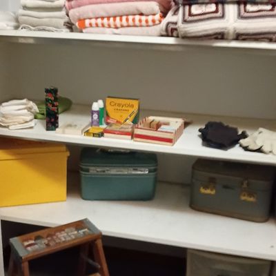 Linen closet shelving filled with folded towels, blankets, crafting supplies, train cases, plastic storage drawers, and a wooden step stool.