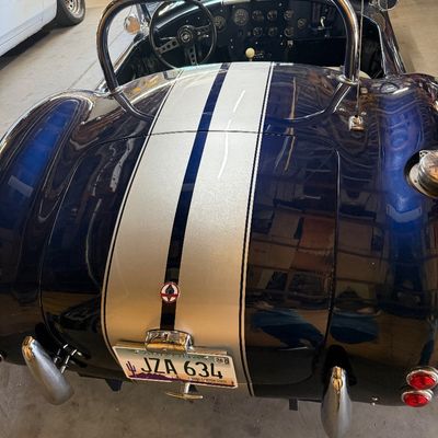 Rear view of a Shelby Cobra replica roadster in dark metallic blue with silver racing stripes, chrome roll bar, and Arizona license plates.