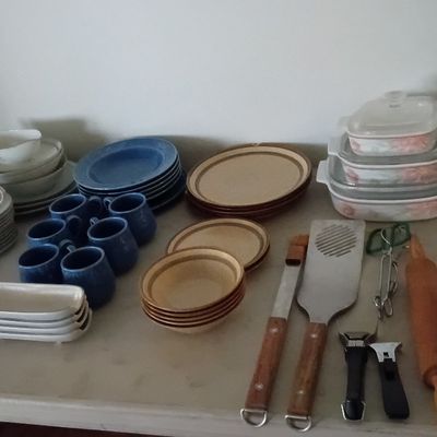 Assortment of vintage kitchenware including CorningWare casserole dishes, stoneware plate and mug sets, and various utensils on a tabletop.