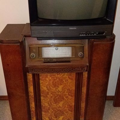 Vintage Art Deco wooden floor-standing console radio featuring patterned fabric speaker grille, topped with a 1990s Toshiba black CRT...