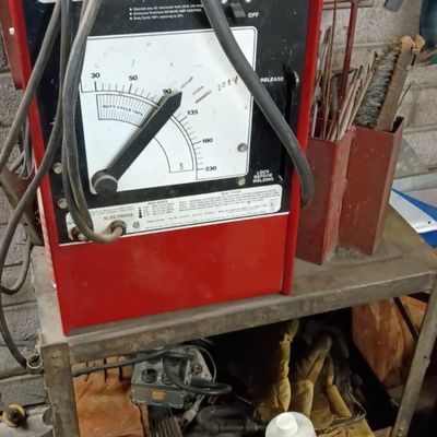Vintage red 230 arc welder mounted on a metal utility cart, accompanied by welding cables, rods, and workshop supplies.