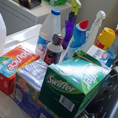 Assorted household cleaning supplies, laundry detergent, and Swiffer products stacked on a washing machine.