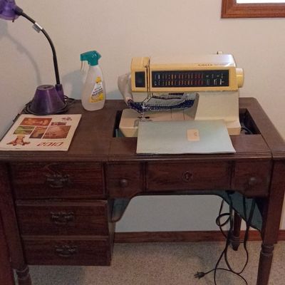 Vintage Singer sewing machine built into a wooden sewing desk with drawers, accompanied by a purple desk lamp.