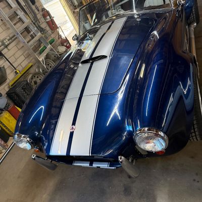 Metallic blue Shelby Cobra replica roadster with silver racing stripes, chrome roll bar, and side pipes.
