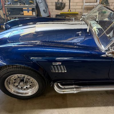 Blue AC Cobra replica roadster featuring silver racing stripes, side exhaust pipes, chrome wheels, and 427 badging.