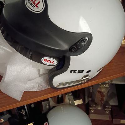 White Bell Mag RS open-face racing helmet with a black sun visor, displayed alongside a second Simpson helmet and racing trophies.