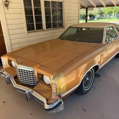 Vintage late-1970s Ford Thunderbird in gold paint with a white vinyl top, parked under a carport.