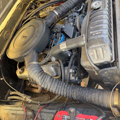 Vintage vehicle engine bay featuring a Ford V8 engine with a round air cleaner assembly, radiator, and a red Optima battery.