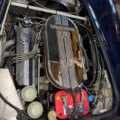 Engine bay of a blue Shelby Cobra replica featuring a chrome oval Cobra air cleaner, finned valve covers, and an Optima red top battery.