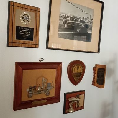 Collection of racing memorabilia, including a framed vintage photograph of a drag racing car, commemorative plaques, and wall awards.