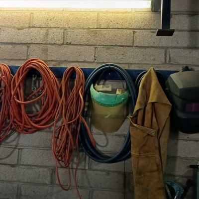 Assorted heavy-duty orange extension cords, welding helmets, face shields, and leather protective gear hanging on a garage wall rack.