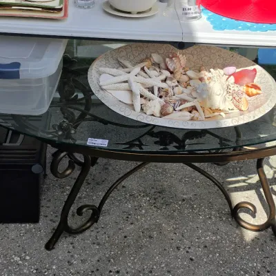 Sale Photo Thumbnail #334: Oval glass-top coffee table with a bronze-toned scrollwork metal base, displaying a large decorative dish filled with sea shells and starfish.