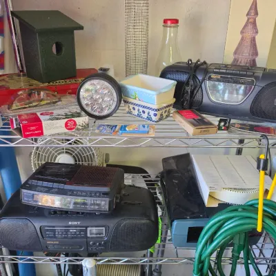 Sale Photo Thumbnail #277: Wire shelving unit filled with vintage audio equipment, including Sanyo and Sony boomboxes, alongside household items, garden supplies, and a green wooden birdhouse.