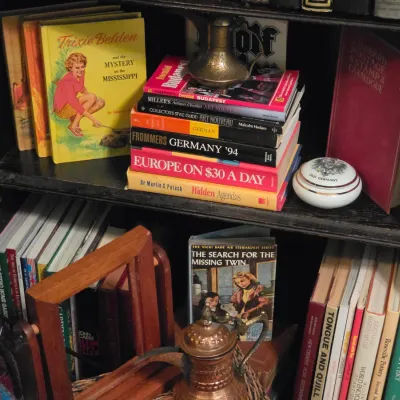 Sale Photo Thumbnail #194: Black wooden bookshelf filled with vintage books, including Trixie Belden and Vicki Barr mystery titles, travel guides, and decorative brass and porcelain items.