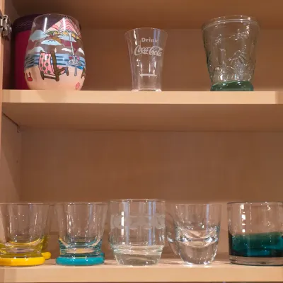 Sale Photo Thumbnail #17: Collection of assorted glassware inside a cabinet, featuring vintage Coca-Cola branded glasses, hand-painted beach scenes, and colorful weighted-base tumbler glasses.