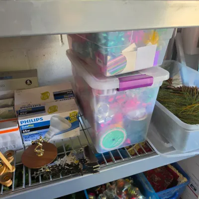 Sale Photo Thumbnail #301: Shelving unit filled with holiday supplies, including clear plastic storage totes of gift bows and greenery, boxed Philips light bulbs, and wooden Christmas decor.