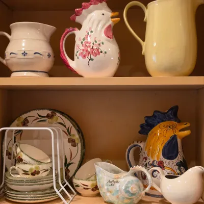 Sale Photo Thumbnail #34: Collection of decorative ceramic pitchers, teacups, and dinnerware inside a wooden cabinet, including hand-painted rooster novelty pitchers.