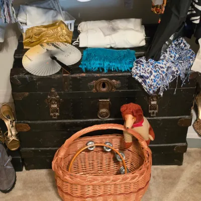 Sale Photo Thumbnail #196: Antique black steamer trunk displaying folded textiles, a folding fan, and surrounded by shoes, a wicker basket, and a vintage stuffed animal.