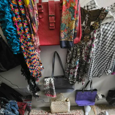 Sale Photo Thumbnail #164: Collection of women's fashion items, including colorful knit scarves, floral dresses, plaid shirts, and various leather and fabric handbags displayed on a wall.